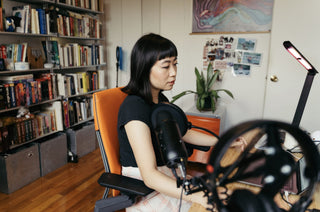 Image of artist Nicole He working at her desk. She has medium length dark hair and is wearing a black tshirt. Behind her is a bookcase